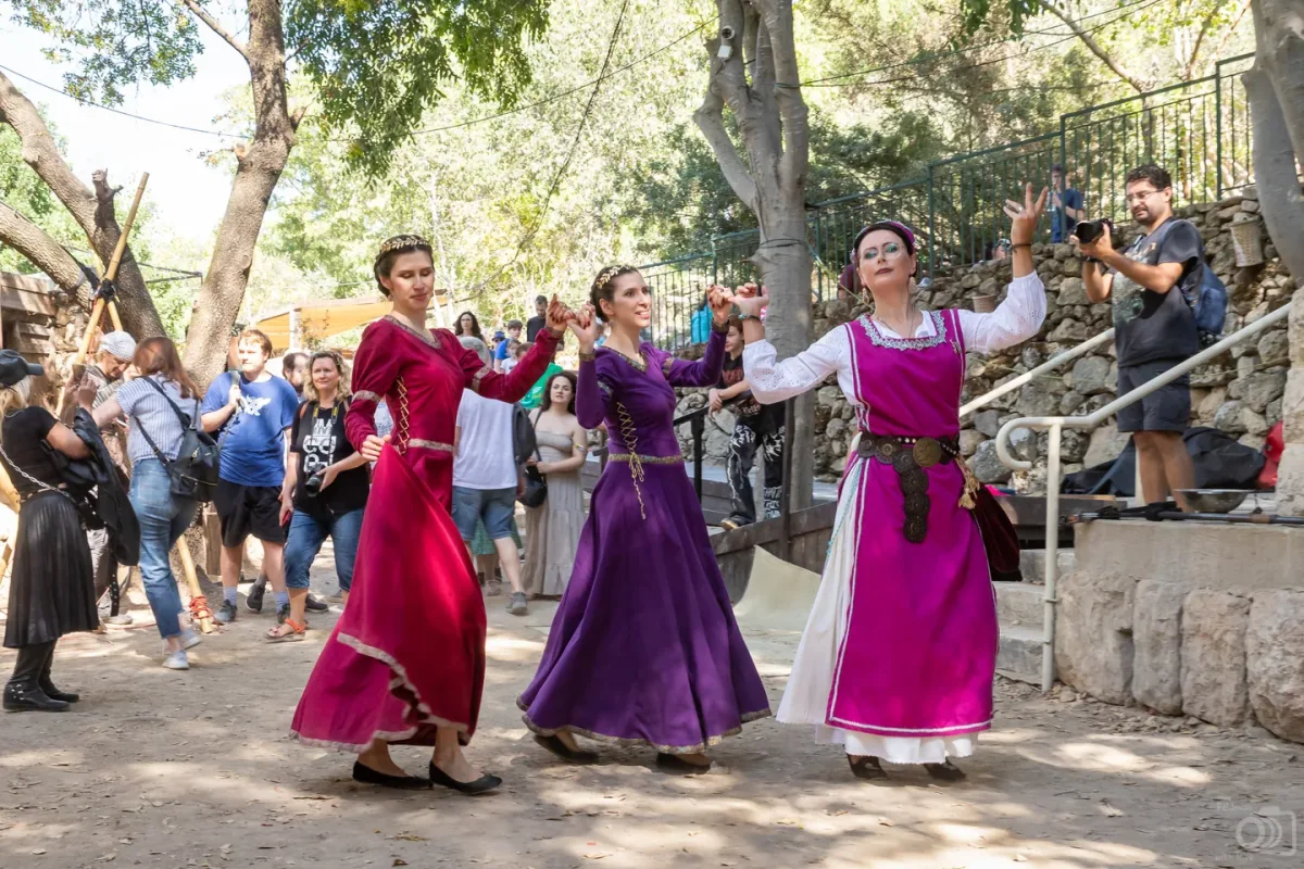 שלוש משתתפות בתלבושות היסטוריות צבעוניות מבצעות ריקוד מימי הביניים.